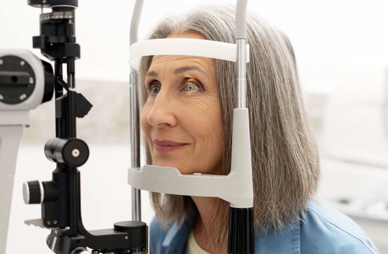 Older woman with gray hair undergoing an eye examination using a slit-lamp machine, looking forward calmly during the test in a bright clinic setting.